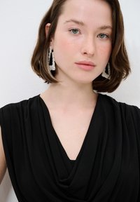 Schwarzes, drapiertes Top mit einem V-Ausschnitt. Das Model hat kurze, wellige Haare. Es trägt große, silberne geometrische Ohrringe mit funkelnden Akzenten.