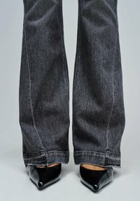 Nogi ubrane w czarne dżinsy z szerokimi nogawkami i surowymi brzegami, zakrywające czarne, szpiczaste szpilki na jasnym tle.