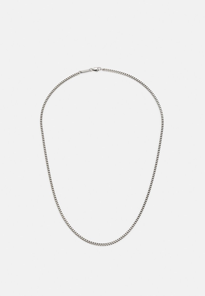 Collier chaîne fine en boîte argentée avec fermoir mousqueton, disposé en forme ovale sur un fond blanc uni.