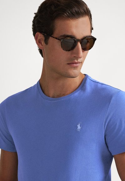 Μπλε κοντομάνικο μπλουζάκι με στρογγυλή λαιμόκοψη και διακριτικό κεντημένο λογότυπο. Μαύρα γυαλιά ηλίου με σκελετό χελώνας αναδεικνύουν την εμφάνιση.