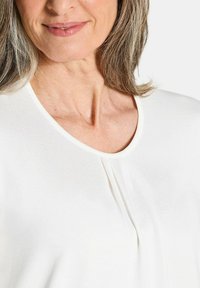 Camicia bianca con scollatura rotonda, caratterizzata da pieghe sottili e una consistenza morbida, completata da una leggera lucentezza e una finitura liscia.