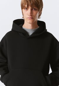 Črna kapucar s kapuco iz mehkega blaga, z sprednjim žepom, sproščenim krojem in rebrastimi manšetami. Gladek tekstura z minimalnimi oblikovnimi detajli.