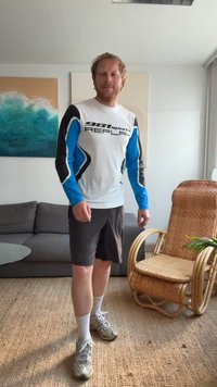 Camisa branca de mangas longas com detalhes em azul e preto, logo "981 sports REPLAY", combinada com calções escuros e ténis claros. Ambiente interno iluminado.