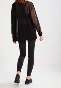 Schwarzes, durchscheinendes Langarmoberteil mit lockerer Passform, kombiniert mit dunklen Leggings und schwarzen Turnschuhen, die ein leichtes, geschichtetes Design zur Schau stellen.