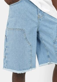 Shorts di denim azzurro chiaro con grandi tasche frontali e orli sfrangiati indossati da una persona con pelle scura e una camicia bianca.