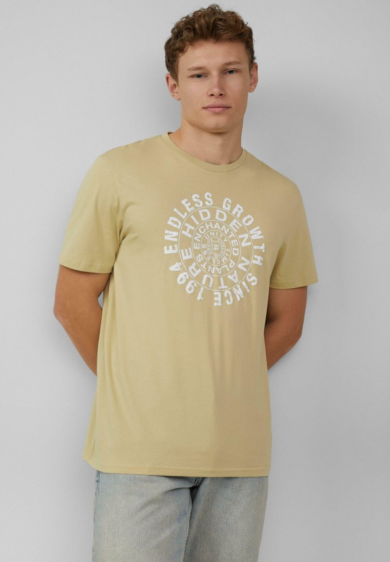 Beige katoenen t-shirt met korte mouwen, ronde hals, met een witte cirkelvormige tekstprint met de tekst "Eindeloze Groei Sinds 1994." Eenvoudig ontwerp.