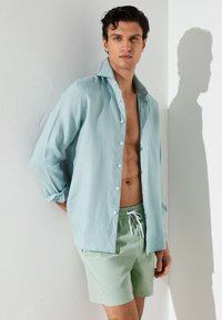 Junger Mann mit dunklem Haar, der ein aufgeknöpftes hellblaues Hemd und mintgrüne Shorts mit Kordelzug trägt, steht vor einer weißen Wand mit Schatten.