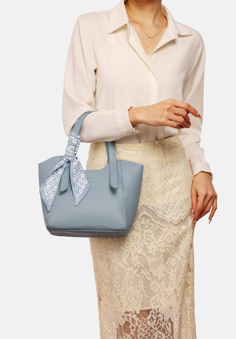 Borsa a tracolla in pelle testurizzata di colore azzurro chiaro con manici corti, impreziosita da un nastro coordinato. Indossata con una blusa crema e una gonna in pizzo.