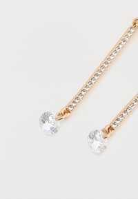 Τα σκουλαρίκια σε χρυσό τόνο διαθέτουν γραμμικό σχέδιο με μια σειρά από καθαρά κρυστάλλινα στοιχεία και δύο μεγαλύτερους στρογγυλούς διαυγείς λίθους στις άκρες.