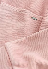 Blød lyserød fløjlstof med et struktureret ribmønster, der har et lille sølvmærke med påskriften "DOWNTIME" syet ind i kanten.