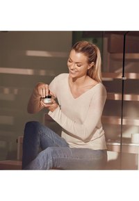 Ljus beige långärmad tröja med V-hals, kombinerad med blå jeans. Personen håller ett litet glas med svart lock, sittande på ett trappsteg.