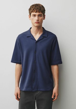 Junger Mann trägt ein navyblaues, kurzärmeliges, strukturiertes Hemd mit Knöpfen und dunkle Hosen, steht vor einem schlichten hellgrauen Hintergrund.