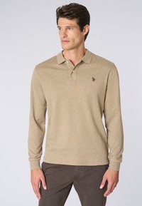 Uomo che indossa una polo a maniche lunghe beige e pantaloni scuri, che guarda di lato su uno sfondo chiaro e semplice.