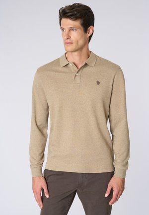 Uomo che indossa una polo a maniche lunghe beige e pantaloni scuri, che guarda di lato su uno sfondo chiaro e semplice.