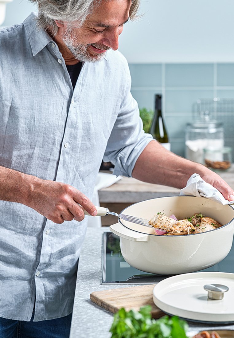 Biały, emaliowany żeliwny garnek z pokrywką, wypełniony ugotowanym kurczakiem i ziołami. Mężczyzna używający widelca, aby nabrać potrawę z garnka na blacie.