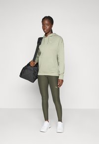 Felpa verde oliva con tasca a marsupio e logo, leggings verdi oliva abbinati, sneakers bianche e borsa a tracolla nera. Tessuto liscio, design casual.