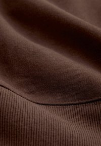 Brauner Stoff mit gerippter Struktur, der glatte Konturen und subtile Nähte aufweist. Das Material wirkt weich und flexibel.