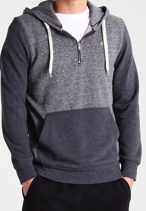 Hombre con una sudadera con capucha de dos tonos de gris, cremallera frontal, cordones blancos y un bolsillo tipo canguro, con las manos en los bolsillos de un pantalón negro.