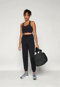 Schwarzer Sport-BH und Jogginghose mit Kordelzug, kombiniert mit grau-weißen Sneakers. Schwarze Tasche mit Griff in der rechten Hand, einfarbiger Hintergrund.