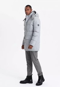 Chaqueta acolchada gris claro con capucha, botones a presión en el frente, bolsillos laterales y un parche de logo. Lleva puesta con pantalones grises y botas negras de cordones.