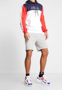 Sudadera con capucha roja y marina con acentos blancos, pantalones cortos grises y zapatillas deportivas multicolores, que presentan un logo en el pecho y en los pantalones cortos.
