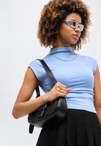 Γυναίκα με σγουρά μαλλιά που φορά ασημένια γυαλιά ηλίου, γαλάζιο αμάνικο τοπ, μαύρη φούστα, ασημένιο κολιέ αλυσίδα και κρατά μαύρη δερμάτινη τσάντα ώμου.