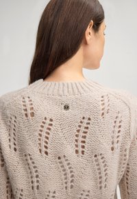 Hellbeige gestrickter Pullover mit einem strukturierten Muster aus offenen Maschen und Rundhals, akzentuiert durch einen kleinen Logo-Knopf auf der Rückseite.