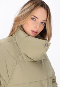 Giacca puffer khaki con colletto alto, design geometrico a trapuntatura e bottoni metallici circolari. Texture morbida e forma slanciata.