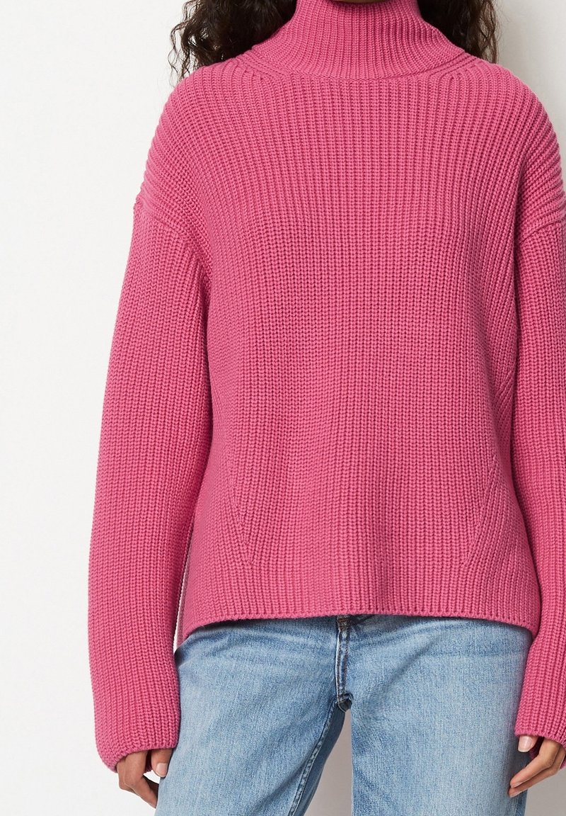 Pull rose en tricot avec un col montant, des manches larges et une texture côtelée. Associé à un jean bleu clair.