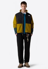 Chaqueta de forro polar con la parte superior negra y los bolsillos inferiores mostaza, llevada sobre una camiseta azul. Pantalones negros y zapatos marrones completan el conjunto. Llaves sujetas.