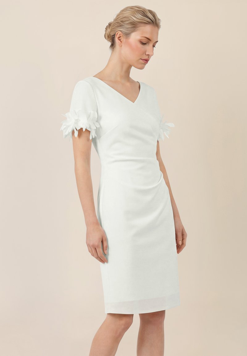 Vrouw met blond haar, gekleed in een witte jurk tot op de knie met een V-hals en korte mouwen, versierd met 3D stoffen bloemdetaillering.