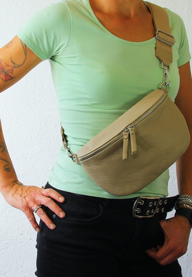 Beige leren schoudertas met een ronde vorm, twee ritsen, en een gestructureerde afwerking. Verstelbare strap met metalen hardware accenten.