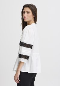 Chemisier blanc avec des manches bouffantes, des accents en dentelle noire et un ourlet peplum ; fabriqué à partir d'un tissu léger, il présente de délicates broderies.