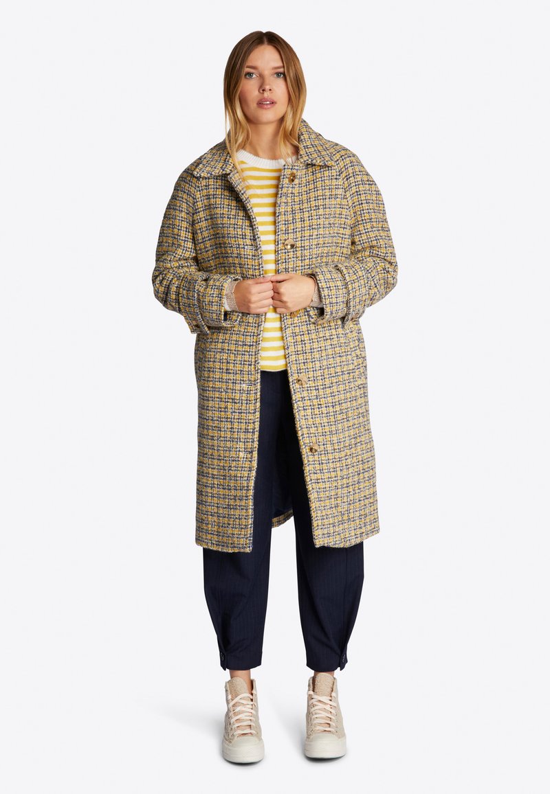 Abrigo de tejido con un patrón a cuadros en amarillo y azul, con un cuello, botones frontales y bolsillos laterales. Llevado sobre un suéter a rayas.