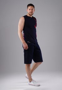 Man wearing a navy sleeveless shirt with red accents, dark denim shorts, and white sneakers standing against a plain gray background.