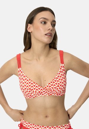 Mujer con un top de bikini rojo y blanco con patrón de zigzag y tirantes trenzados, mirando ligeramente hacia su derecha con las manos en las caderas.