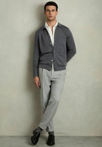 Grå cardigan med krave-design, knaplukning fortil og brystlomme, parret med lysegrå skræddersyede bukser og sorte sko.