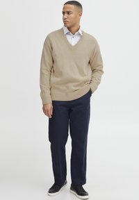 Beige V-Ausschnitt Pullover aus weichem Material, getragen über einem hell gestreiften Hemd, kombiniert mit navyfarbener Hose und schwarzen Sneakers.