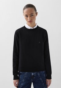 Sudadera negra de mezcla de algodón, con un cuello redondo, puños y dobladillo acanalados. Pequeño detalle de logo en el pecho, combinada con jeans de mezclilla.