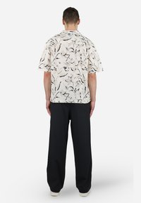 Kortsleeve, lysebeige skjorte med et svart og kremfarget abstrakt mønster; kombinert med løstsittende svarte bukser og hvite joggesko.
