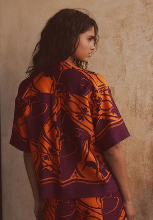 Žena s vlnitými tmavými vlasmi, ktorá má na sebe voľnú oranžovú a tmavofialovú vzorovanú košeľu s krátkym rukávom a zodpovedajúce nohavice pred textúrovanou béžovou stenou.