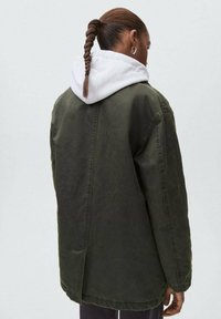 Ylikoko vaakavihreä takki, jossa on rakennettu pinta, ja valkoinen huppu kurkistelee ylhäältä, sekä suora takisauma.