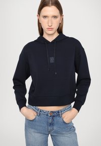 Navyvärinen lyhythihainen huppu, jossa on teksturoitu pinta, kenguru-tasku, kiristysnauha huppua varten ja hillitty logo-patch etupuolella.