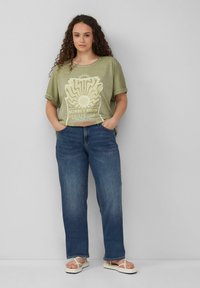 Olivgrünes T-Shirt mit kurzen Ärmeln und weißem Grafikdruck, kombiniert mit blauen Jeans im geraden Schnitt und weißen Flip-Flops. Einfaches Design und lässige Passform.
