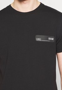 Svart bomulls t-shirt med rund halsringning och en rektangulär logotyppatch på vänster bröst, med vit text och detaljer.