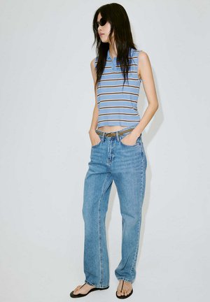 Femme portant des lunettes de soleil noires, un haut rayé bleu sans manches, un jean bleu clair et des tongs noires, debout les mains dans les poches.