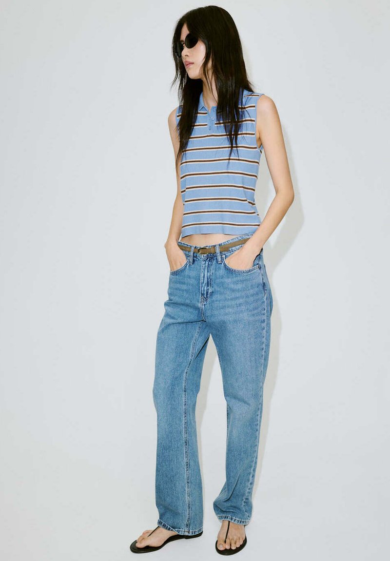 Femme portant des lunettes de soleil noires, un haut rayé bleu sans manches, un jean bleu clair et des tongs noires, debout les mains dans les poches.