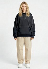 Felpa con cappuccio nera in cotone con tasca frontale e cappuccio con coulisse, abbinata a pantaloni beige larghi e sneakers bianche.