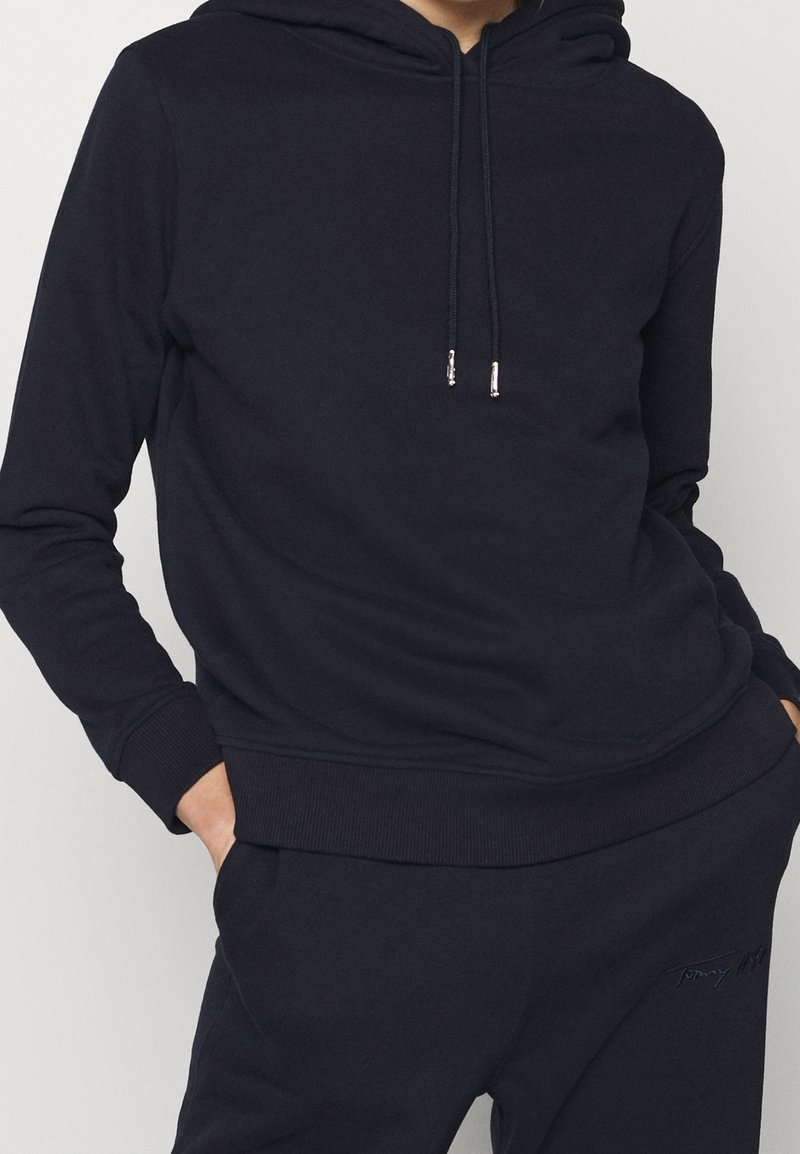 Sudadera con capucha azul marino hecha de material suave, con bolsillo tipo canguro, cordones con puntas metálicas y puños y dobladillo acanalados.