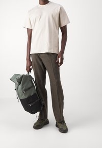 Mies, který nosí béžovou košili s krátkým rukávem, hnědé pruhované kalhoty, zelené tenisky a drží zelený a černý batoh v ruce na bílém pozadí.